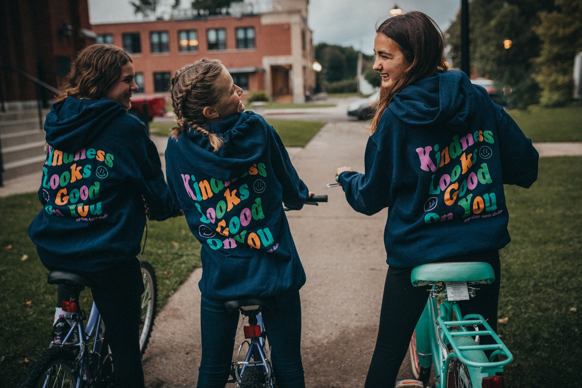 Three Signature Dance students on bikes wearing navy 'Kindness Looks Good on You' hoodies